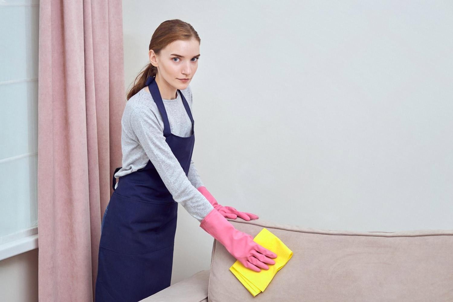 Young woman in an apron wiping down a beige upholstered sofa with a yellow cloth